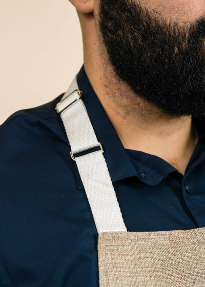closeup of a white apron strap on a man's shoulder, including part of the apron and the man's bearded face