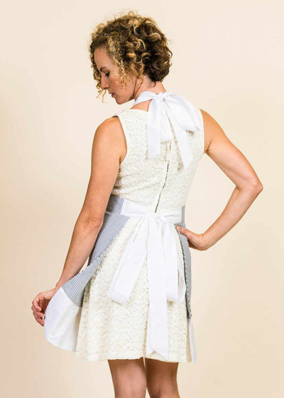 blonde woman wearing a white dress, with her back to the camera.  The corner of her white seersucker apron with white ruffle is visible