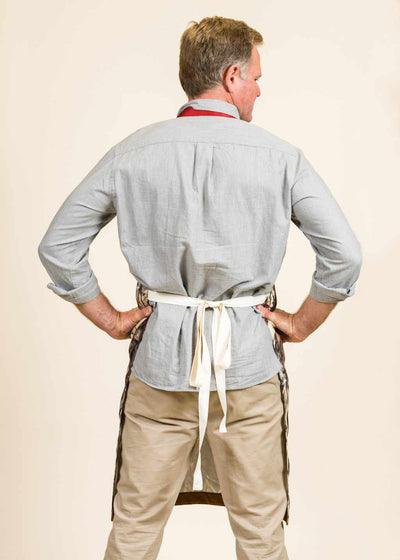 The backside of a man wearing a blue shirt, khaki pants, and apron with white straps around his back.