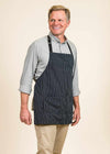 side front view of smiling man wearing a black apron with white stripes and a light blue shirt. The man is blond